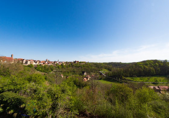 Blick über das grüne Taubertal mit Wiesen, Bäumen und der Altstadt von Rothenburg ob der Tauber im Hintergrund unter blauem Himmel